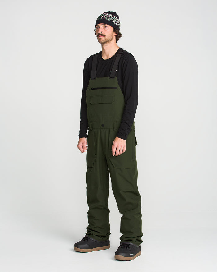 A man stands against a plain white background wearing a black long-sleeve shirt, Volcom Men’s Roan Bib crafted from recycled stretch Oxford for enhanced comfort and mobility, black shoes, and a patterned beanie. The dark green Volcom overalls feature 15K protection, delivering both style and functionality.