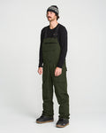 A man stands against a plain white background wearing a black long-sleeve shirt, Volcom Men’s Roan Bib crafted from recycled stretch Oxford for enhanced comfort and mobility, black shoes, and a patterned beanie. The dark green Volcom overalls feature 15K protection, delivering both style and functionality.