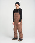 A man with a mustache stands facing forward against a plain white background, wearing a black beanie, a black long-sleeve shirt, and brown Men’s Roan Bib overalls by Volcom, crafted from recycled stretch Oxford. He is also wearing black shoes.