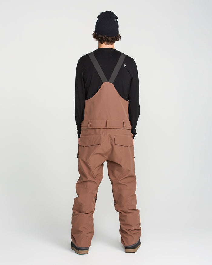 A person wearing Volcom Men’s Roan Bib snow pants in brown, crafted from recycled stretch Oxford fabric and paired with black suspenders and a black long-sleeve shirt, is facing away from the camera. They stand against a plain white background, ready for winter weather with 15K protection.