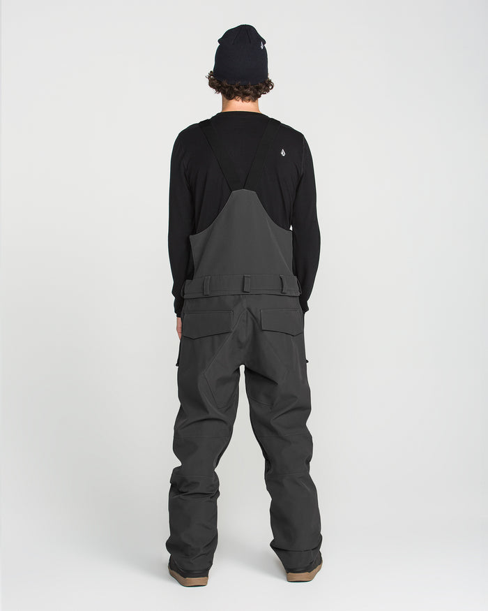 A person stands facing away, wearing a black beanie, a black long-sleeve shirt, and the Volcom Men’s Roan Bib in dark gray—featuring multiple pockets and snow pant styling—against a plain white background.