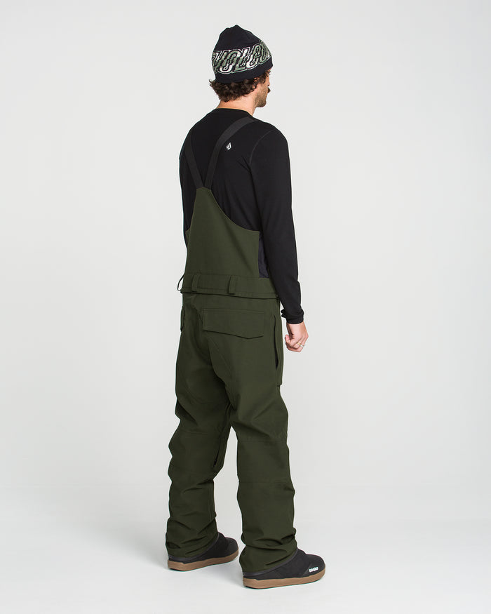 A person stands facing away, wearing a black long-sleeve shirt and the Volcom Men’s Roan Bib in olive green—expertly crafted from recycled stretch Oxford for comfort and durability—paired with black shoes and a black beanie with white writing, all set against a plain white background.