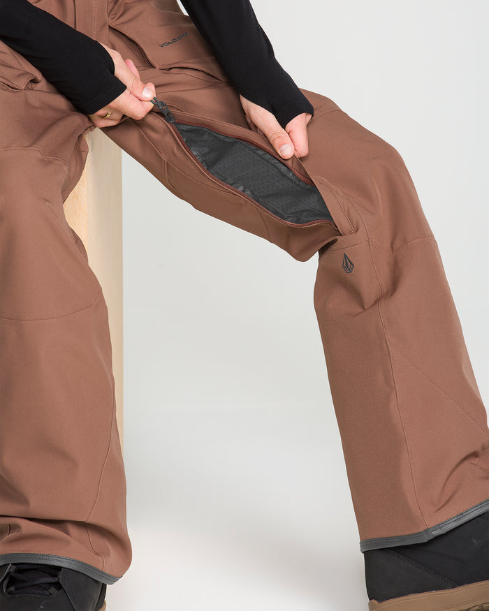 A person wearing Volcom Men’s Roan Bib pants in brown sits on a light wooden stool, unzipping the ventilation panel on the side of their recycled stretch Oxford pants. Only their lower body and hands are visible.
