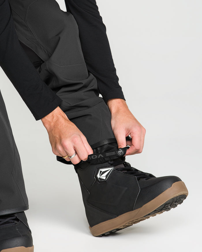 A person wearing Volcom Men’s Roan Bib fastens the strap on a black snowboard boot with a brown sole. The background is light and uncluttered.