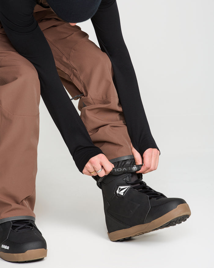 A person wearing Volcom Men’s Roan Bib pants in brown—crafted from recycled stretch Oxford fabric for 15K weather protection—and black snow boots is tightening the strap on one boot. The image features only the lower half of their body against a plain white background, emphasizing the performance gear.