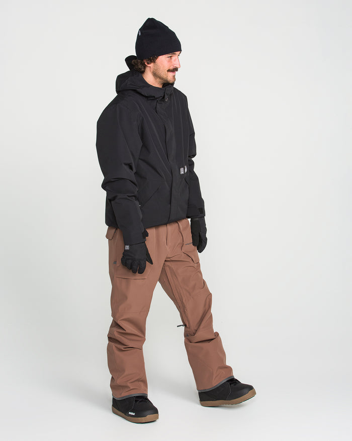 A person wearing a black beanie, black jacket, black gloves, brown pants—reminiscent of the Volcom Men’s Roan Bib—and black boots stands against a plain white background, looking to the side with a slight smile.
