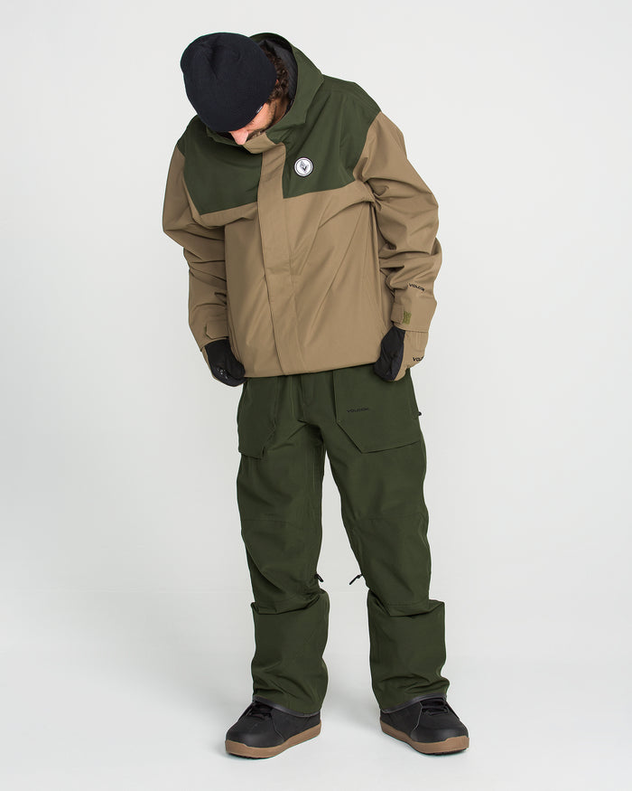 A person is wearing the Volcom Men’s Roan Bib—a pair of green bib pants—paired with a tan and green jacket made from recycled stretch Oxford fabric with 15K protection, along with black gloves, a black beanie, and black shoes. The individual is standing indoors against a plain white background and looking downward.