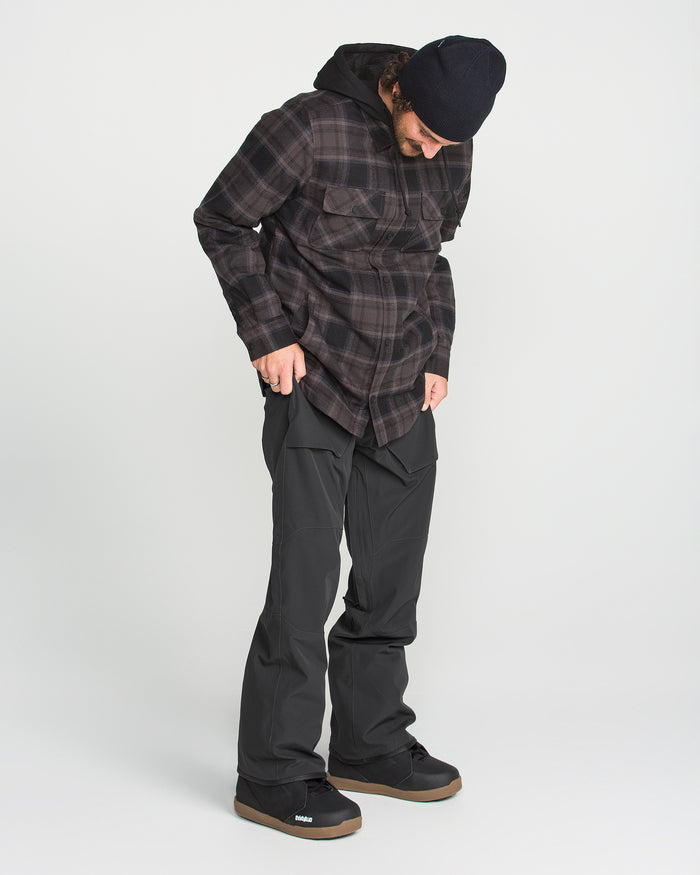 Wearing the Volcom Men’s Roan Bib, a person in a black beanie and plaid shirt stands on a white background, looking down.