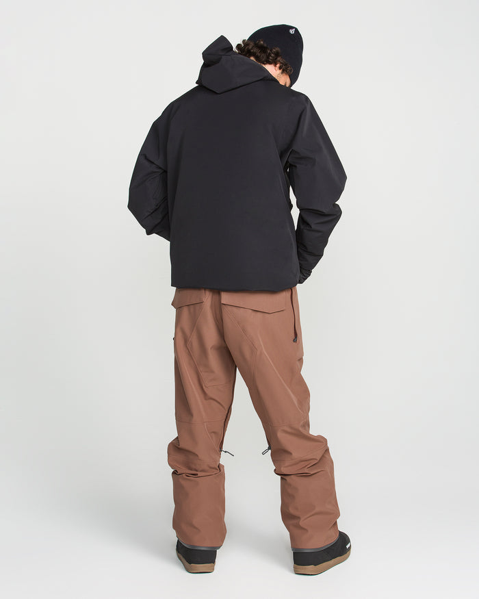 A person stands with their back to the camera against a plain white background, wearing Volcom’s Men’s Roan Bib brown overalls, a black hooded jacket made from recycled stretch Oxford with 15K weather protection, black boots, and a black beanie. Their hands are tucked into the jacket pockets, highlighting its reliable weather resistance.