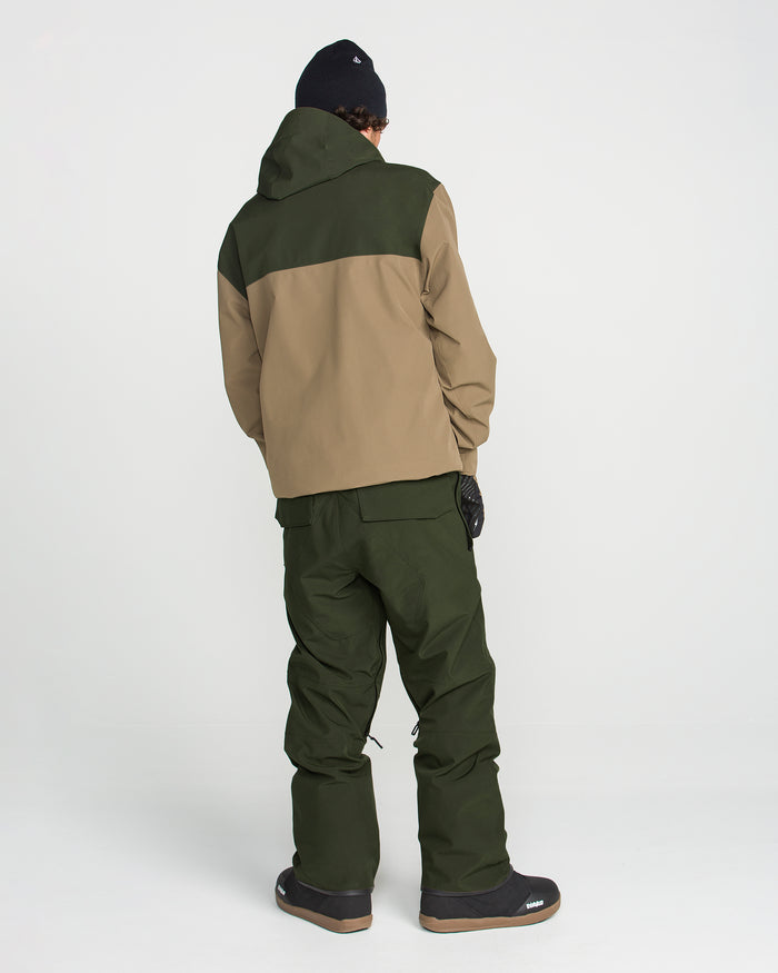 A person stands with their back to the camera against a plain white background, wearing Volcom Men’s Roan Bib snow pants in green, paired with a two-tone green and tan recycled stretch Oxford jacket, black gloves, a black beanie, and black winter boots.