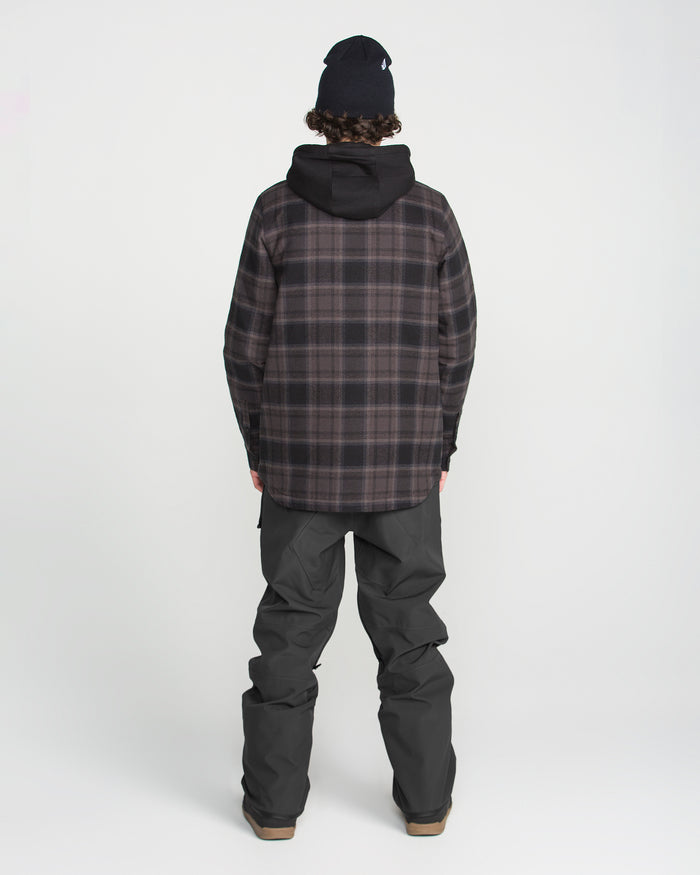 A person stands facing away from the camera against a plain white background, wearing the Volcom Men’s Roan Bib snow pants, paired with a plaid jacket and black beanie.