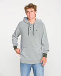 A young man wearing the Volcom Men's Rowan Thermal Long Sleeve Shirt in light gray, featuring black drawstrings, pairs it with blue jeans as he stands against a plain white background, looking at the camera with a relaxed expression.