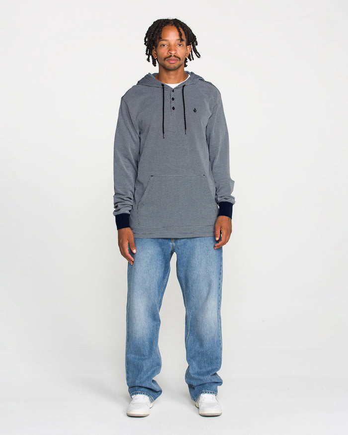 A man with short dreadlocks stands against a plain white background, wearing the Volcom Men's Rowan Thermal Long Sleeve Shirt for midweight warmth, paired with light blue jeans and white sneakers. He looks directly at the camera with a neutral expression.