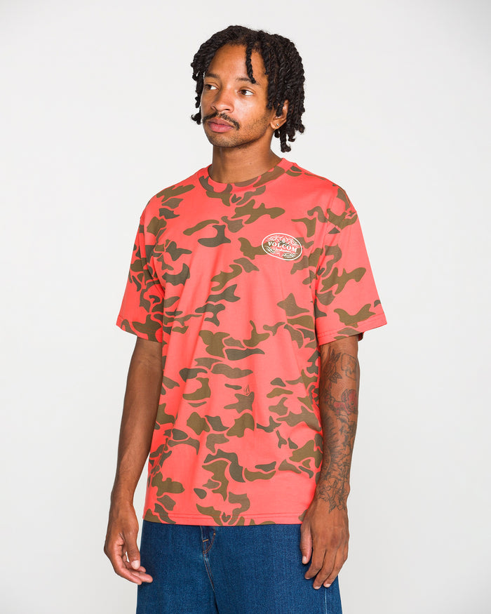 A man wearing the Volcom Men's Safety Workwear Short Sleeve Tee, crafted from premium cotton t-shirt fabric in a pink and olive green camo pattern with a small logo on the chest, paired with blue jeans and standing against a plain white background.