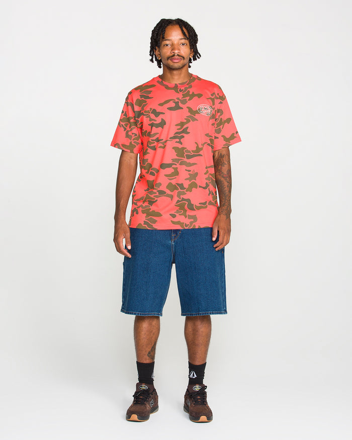 A man stands against a white background wearing the Volcom Men's Safety Workwear Short Sleeve Tee, which features a red camo design and is crafted from premium cotton. He pairs it with blue knee-length denim shorts, black socks, and brown shoes. His short dreadlocks frame his face as he faces forward with a neutral expression.