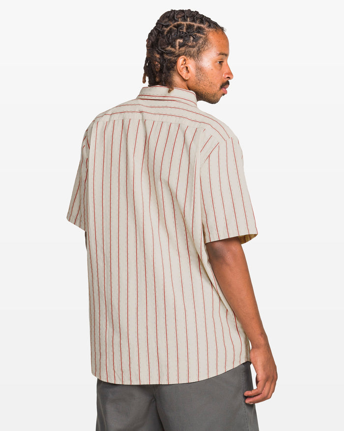 A man with braided hair wears the Volcom Men's Sato Stripe Short Sleeve Tee and gray classic-fit pants. He stands turned away, looking over his shoulder against a plain white background.