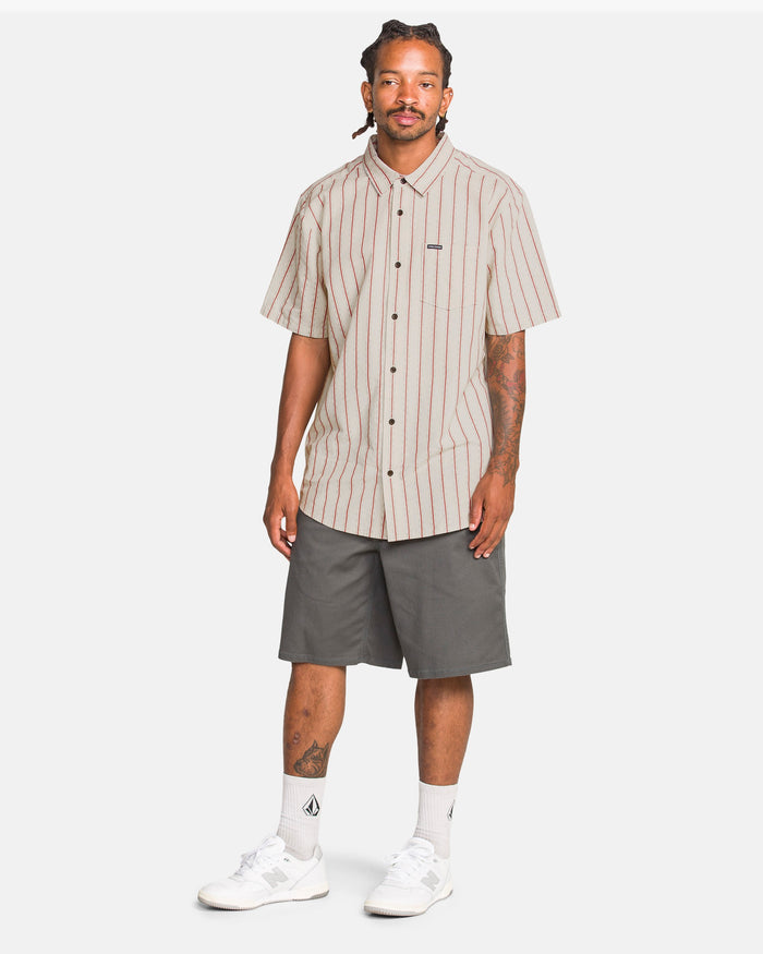 A man with braids and arm tattoos stands facing forward in the Volcom Men's Sato Stripe Short Sleeve Tee, paired with gray knee-length shorts, white crew socks, and sneakers.
