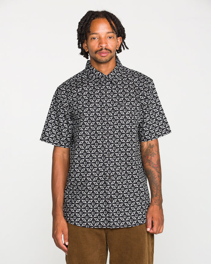 A man with dreadlocks wears the Volcom Men's Scaler Stone Short Sleeve Shirt, featuring a classic fit blackout print with a white geometric pattern, paired with brown pants and standing against a plain white background.