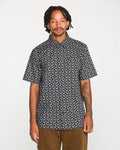 A man with dreadlocks wears the Volcom Men's Scaler Stone Short Sleeve Shirt, featuring a classic fit blackout print with a white geometric pattern, paired with brown pants and standing against a plain white background.