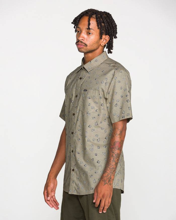 A man with short dreadlocks and tattoos on his left arm wears the Volcom Men's Scaler Stone Short Sleeve Shirt—a light brown, patterned, classic-fit button-up made from a cotton-viscose blend—paired with dark pants, standing against a plain white background.