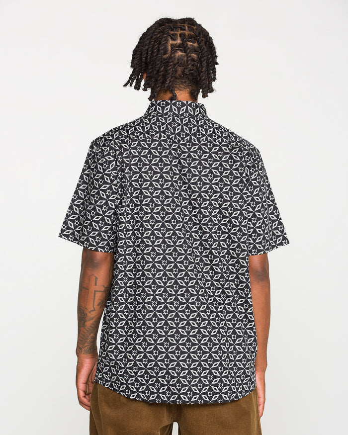 A person with braided hair, seen from behind, wears the Volcom Men's Scaler Stone Short Sleeve Shirt in a blackout print and classic fit, paired with brown pants against a plain white background.
