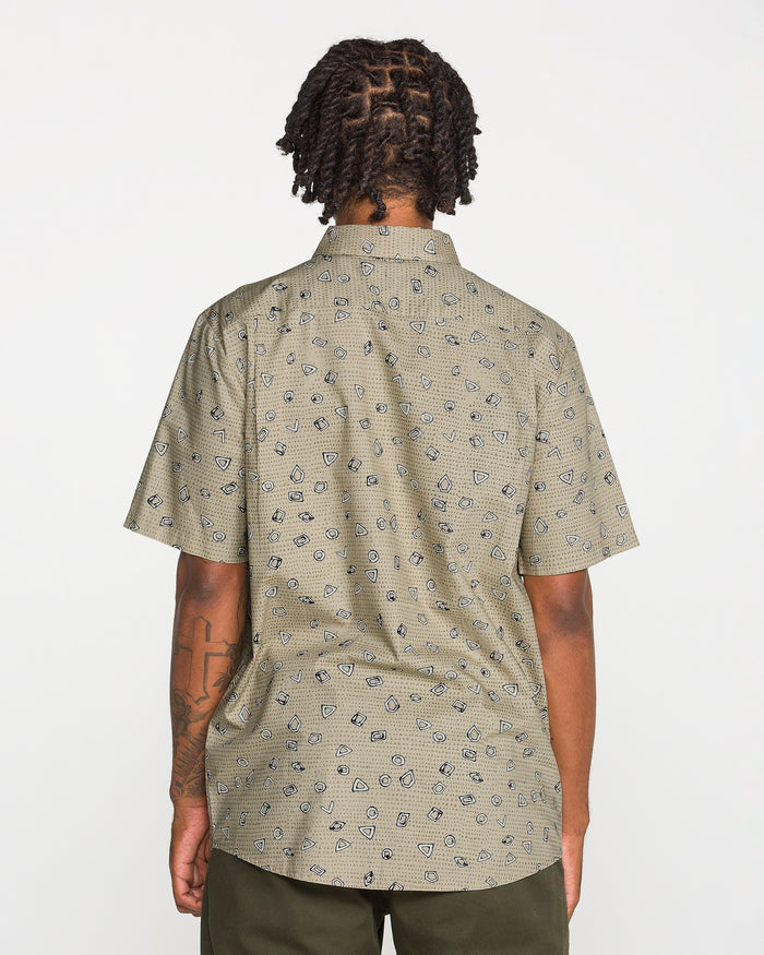 A person with short, twisted hair is photographed from the back against a plain white background wearing Volcom's Men's Scaler Stone Short Sleeve Shirt, featuring a classic fit and beige pattern, paired with olive green pants.