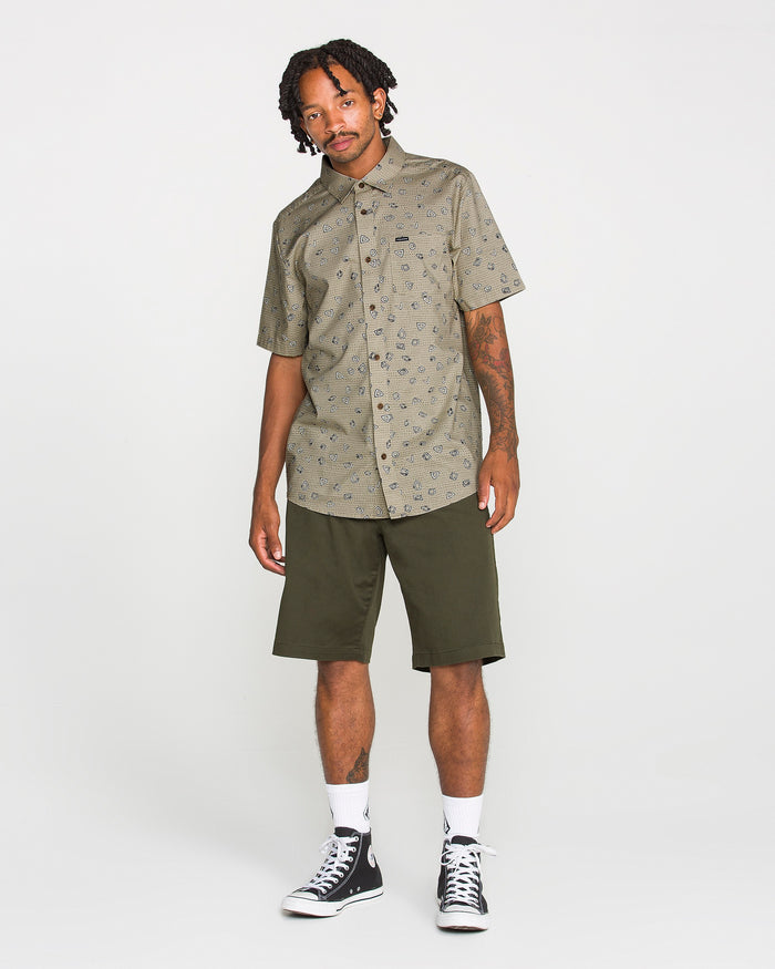 A man stands against a white background, wearing the Volcom Men's Scaler Stone Short Sleeve Shirt featuring an eye-catching pattern, paired with olive green knee-length shorts, white socks, and black high-top sneakers. He has short dreadlocks and visible tattoos on one arm.