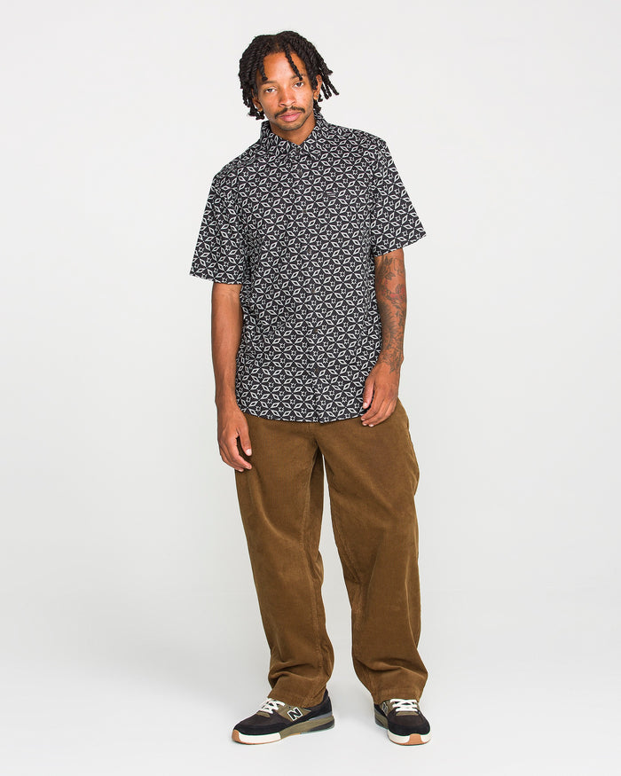 A man with short dreadlocks and visible tattoos on his left forearm stands against a plain white background, wearing the Volcom Men's Scaler Stone Short Sleeve Shirt, brown corduroy pants, and black sneakers.