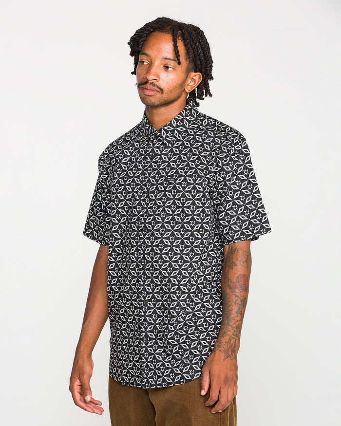 A man with short dreadlocks and tattoos on his arm wears the Volcom Men's Scaler Stone Short Sleeve Shirt and brown pants, standing against a plain white background.