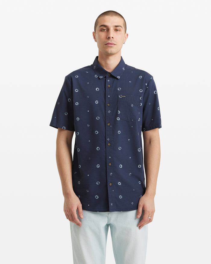 A person stands facing forward against a plain white background, wearing the Volcom Men's Scaler Stone Woven Short Sleeve Shirt—an allover circular dot print shirt in navy blue—with light-colored pants.
