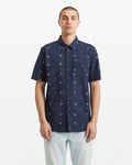 A person stands facing forward against a plain white background, wearing the Volcom Men's Scaler Stone Woven Short Sleeve Shirt—an allover circular dot print shirt in navy blue—with light-colored pants.