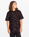 A man with long wavy hair wears the Volcom Men's Scaler Stone Woven Short Sleeve Shirt, a black button-up with red and gray floral print in a classic fit, standing against a plain white background and looking to the side.