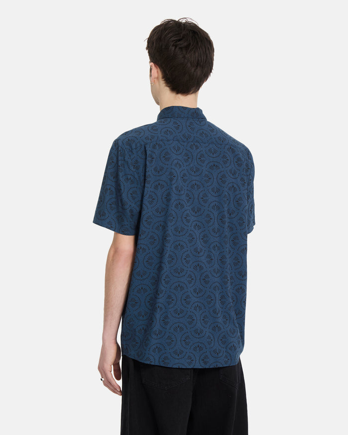 A person with short brown hair stands with their back to the camera, wearing a Volcom Men's Scaler Stone Woven Short Sleeve Shirt in midnight blue and black pants against a plain white background.
