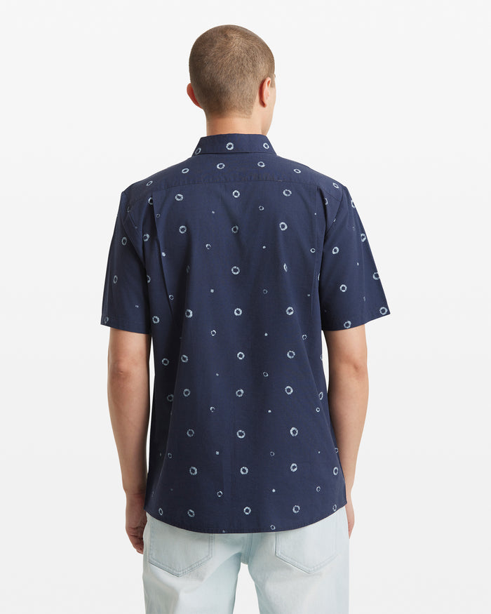 A person is seen from the back wearing a Volcom Men's Scaler Stone Woven Short Sleeve Shirt in navy with light blue circular patterns, paired with light blue jeans against a plain white background.