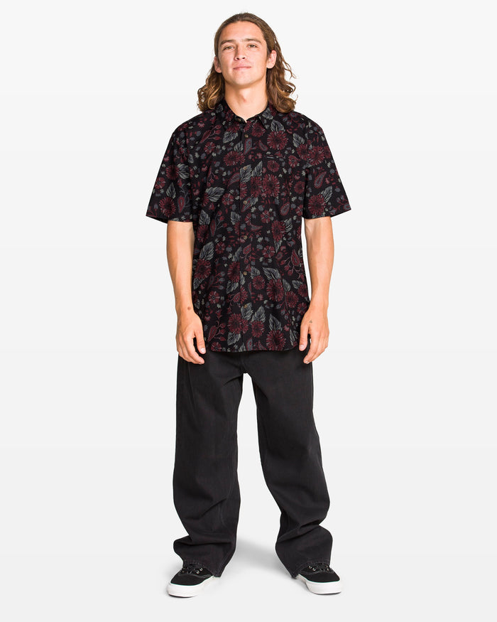 A young person with long hair faces forward, wearing a Volcom Men's Scaler Stone Woven Short Sleeve Shirt in black with red floral patterns, loose black pants, and black sneakers, all set against a plain white background.