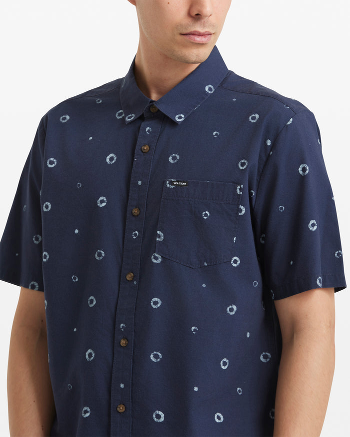 A person is wearing the Volcom Men's Scaler Stone Woven Short Sleeve Shirt in navy blue, featuring an allover circular dot pattern and a chest pocket, while facing slightly to the side.