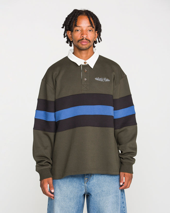 A man stands facing forward against a plain white background, wearing Volcom's Men's Scratch Stone Fleece Polo in dark green with black and blue stripes, a white collar, and light blue jeans. This stylish polo showcases a boxy fit and is crafted from recycled polyester fleece for enhanced comfort and sustainability.
