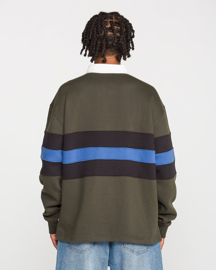 A person with short twists stands facing away, wearing the Volcom Men's Scratch Stone Fleece Polo in a boxy fit olive green with black and blue horizontal stripes, crafted from recycled polyester fleece, paired with light blue jeans against a plain white background.