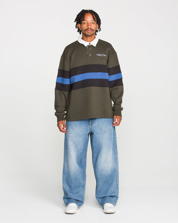 A man stands facing forward against a plain white background, wearing the Volcom Men's Scratch Stone Fleece Polo—featuring a boxy fit in dark green with blue and black stripes—paired with baggy light blue jeans and white sneakers.