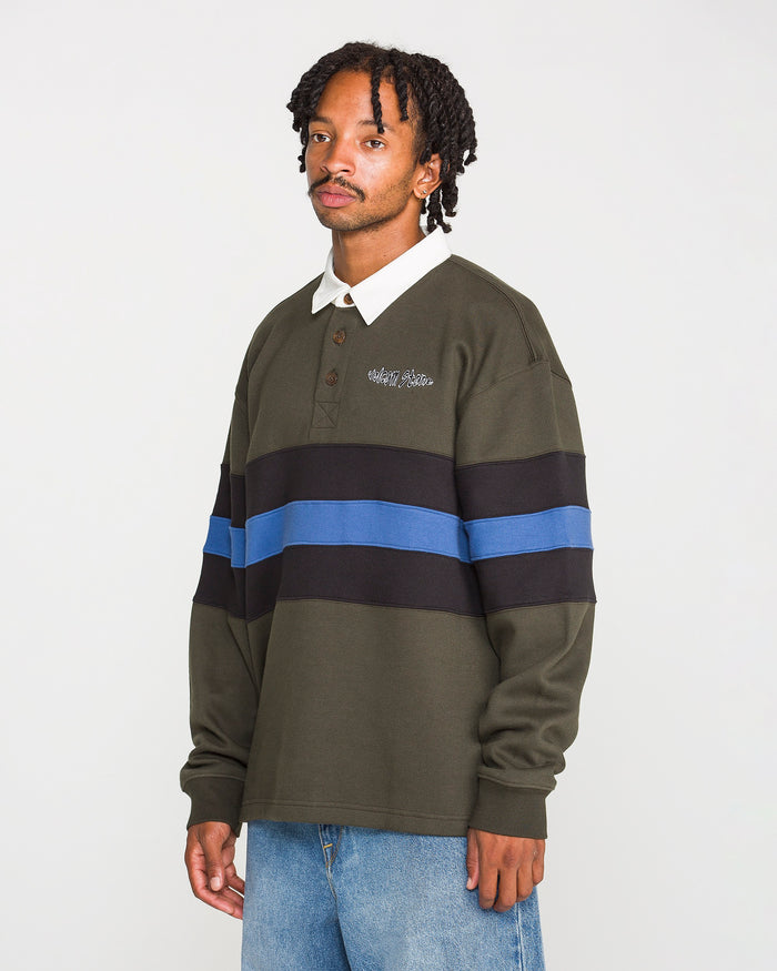 A person wearing the Volcom Men's Scratch Stone Fleece Polo—a dark green long-sleeve shirt with black and blue horizontal stripes in a boxy fit—and light blue jeans stands against a plain white background.