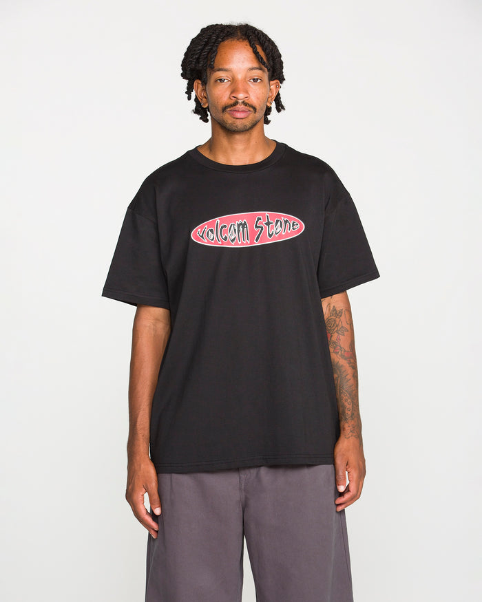 A person with short dreadlocks wears the Volcom Men's Scratch Stone Short Sleeve Tee, featuring a black colorway and a 90's inspired logo, paired with gray pants and set against a plain white background. This loose fit T-shirt is crafted from heavyweight ringspun cotton for exceptional comfort and durability.