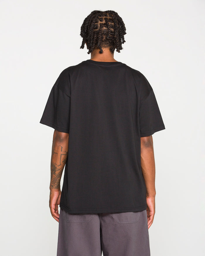 Back view of a person with dark braided hair wearing the Volcom Men's Scratch Stone Short Sleeve Tee in plain black and grey shorts. A large cross tattoo is visible on their right forearm. They are standing against a plain, light grey background.