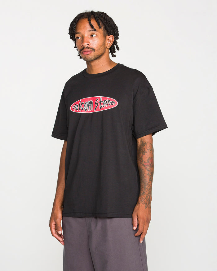 A person wearing the Volcom Men's Scratch Stone Short Sleeve Tee in black, featuring a 90's inspired logo, pairs it with gray pants while standing against a plain white background and looking slightly to the side with one arm relaxed by their side. This loose fit t-shirt is crafted from heavyweight ringspun cotton for comfort and durability.