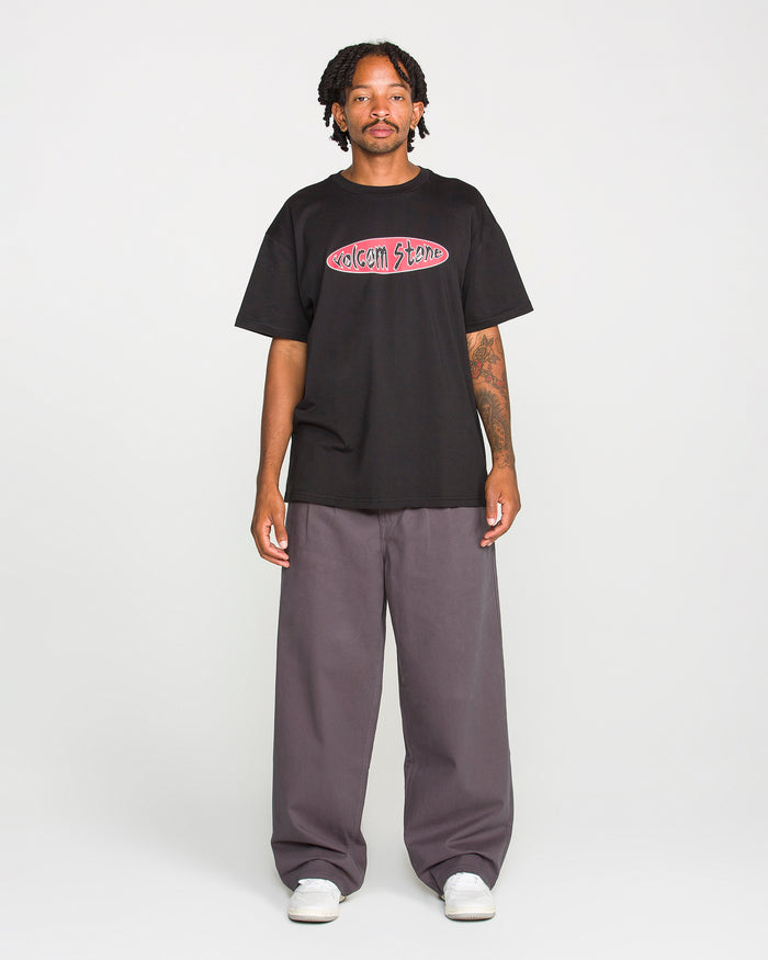 A person stands against a plain background, wearing the Volcom Men's Scratch Stone Short Sleeve Tee in black, made from heavyweight ringspun cotton. They pair it with wide gray pants and white shoes. The individual has short dreadlocks and visible tattoos on one arm.