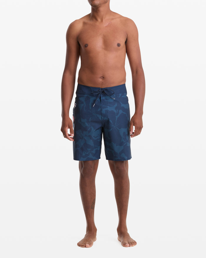 A man stands barefoot against a white background, wearing Volcom Men's Shoreline Mod 20 dark blue patterned board shorts with no shirt. His arms are relaxed at his sides as he faces forward.