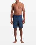 A man stands barefoot against a white background, wearing Volcom Men's Shoreline Mod 20 dark blue patterned board shorts with no shirt. His arms are relaxed at his sides as he faces forward.