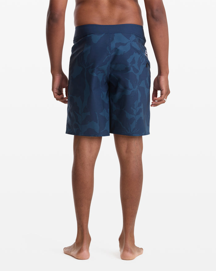 A person stands barefoot facing away, wearing Volcom Men's Shoreline Mod 20 dark blue camo-patterned shorts against a plain white background.