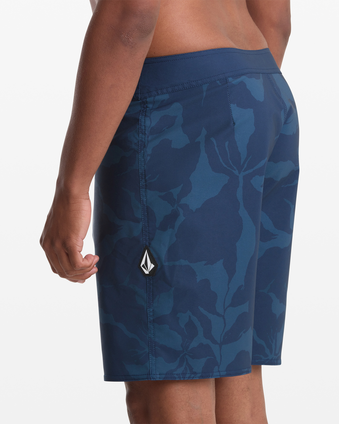 A man models Volcom Men's Shoreline Mod 20 board shorts with a blue pattern and a white and black logo patch on the left leg, shown from the side and back against a plain white background.