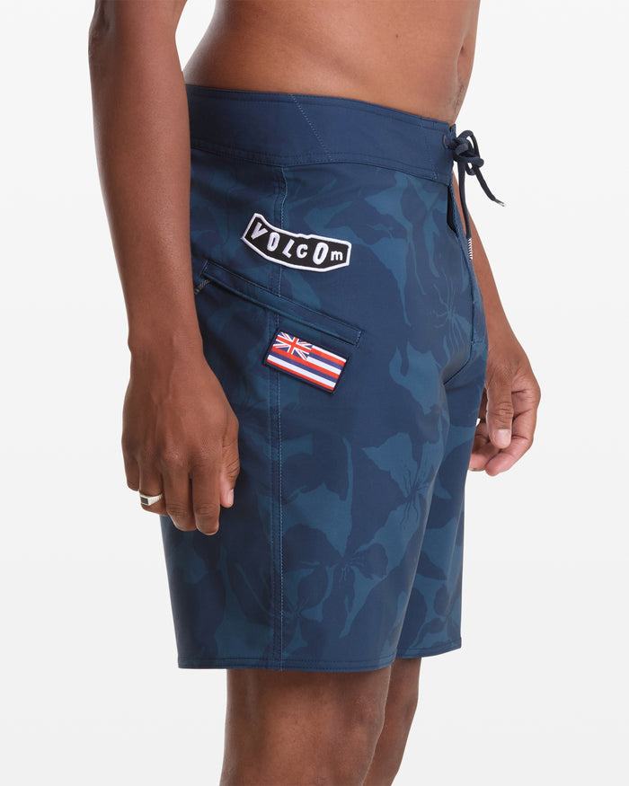 A person wearing Volcom Men's Shoreline Mod 20 board shorts in dark blue floral, featuring a drawstring waist, side pocket, and Volcom patch with a small flag patch on the left side.