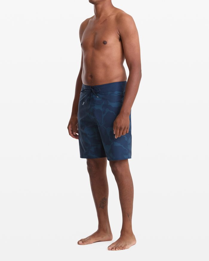 A man stands barefoot against a white background, wearing Volcom Men's Shoreline Mod 20 blue camouflage board shorts. He's shirtless, facing slightly left, with his arms relaxed at his sides.